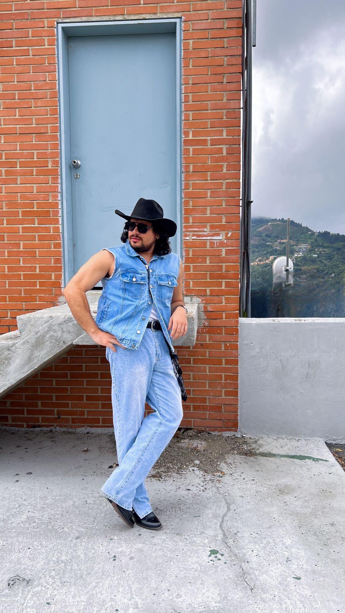 Yisus con sombrero negro, chaleco y pantalón de denim claro, apoyado en un muro de ladrillo
