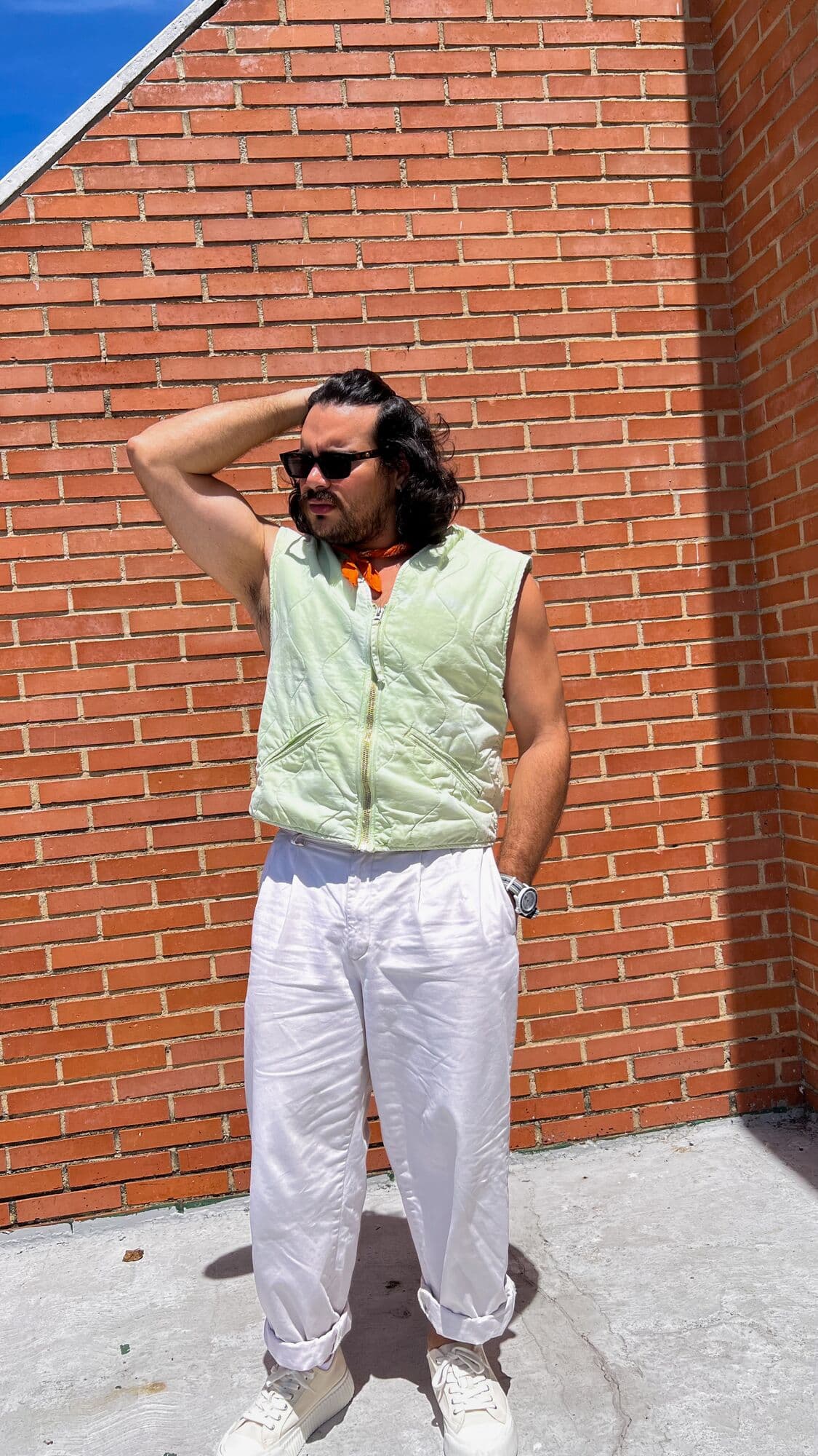 Yisus con chaleco verde menta y pantalón blanco, con una mano en el cabello, frente a ladrillo